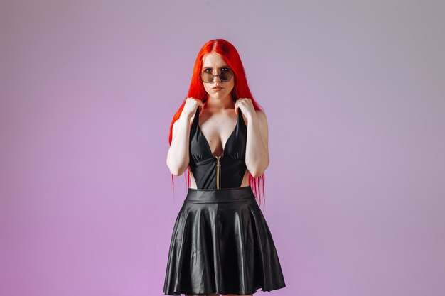 Girl with red long hair in round glasses leather skirt on pink background