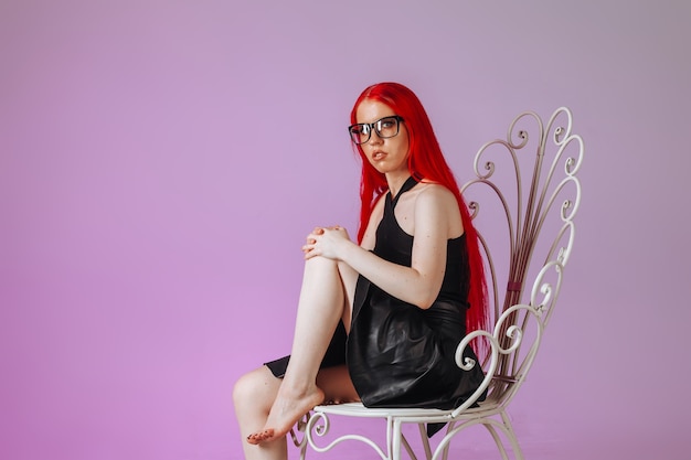 Девушка с рыжими длинными волосами в очках и кожаной юбке сидит на ажурном стуле