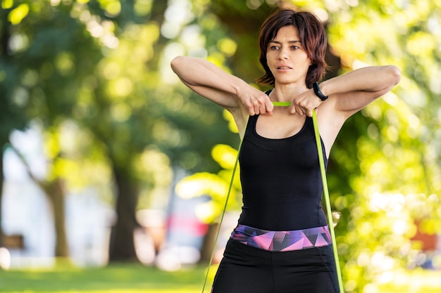Девушка с резиновой резинкой тренируется для рук с сопротивлением на открытом воздухе в парке бывшая молодая женщина