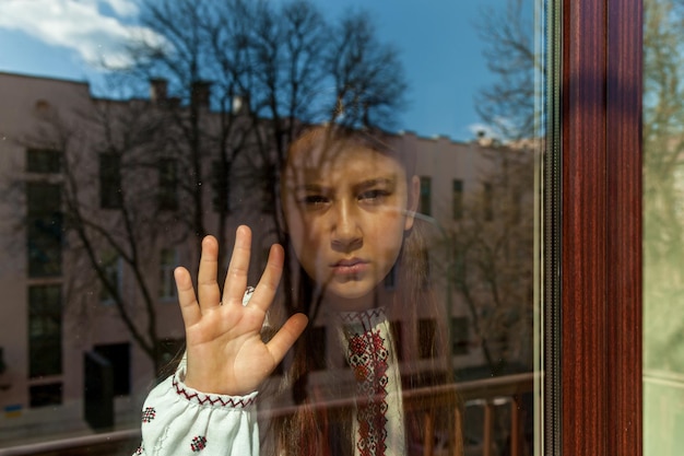 A girl with a sad face in an embroidered dress stands near the window The girl protests against