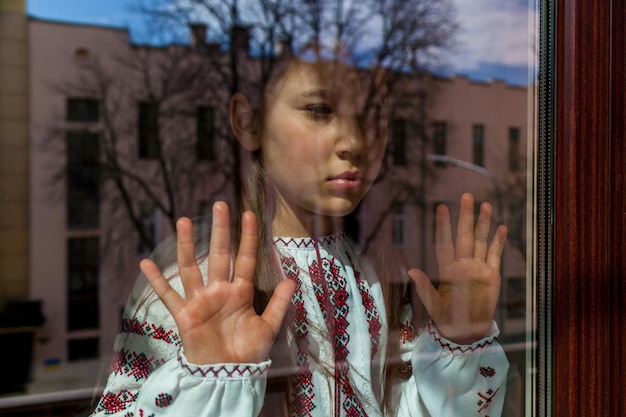 A girl with a sad face in an embroidered dress stands near the window The girl protests against