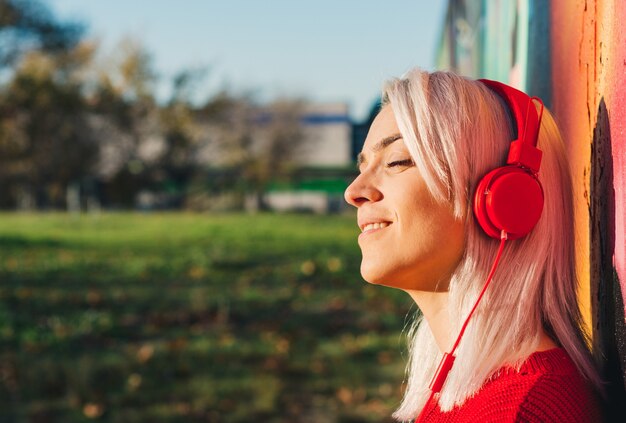 Girl with silver hair listening to music with red headphones.
leaning on a graffiti wall.