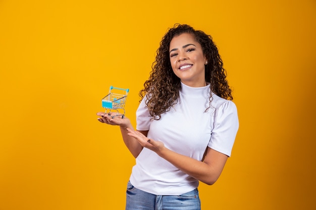 Ragazza con la piccola carta della spesa. donna sorridente e ballante dei capelli ricci nel concetto di acquisto. giovane donna con un carrello in miniatura. e-commerce e affari. macchina della spesa. acquirente donna. sfondo giallo.