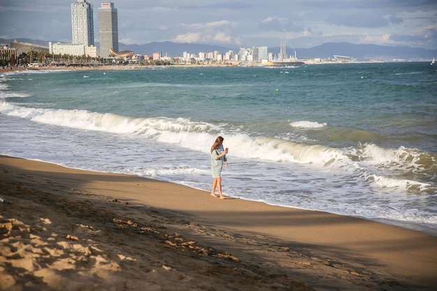 Девушка со смартфоном стоит на пляже в солнечный день Стильный хипстер у волн на море Женщина разговаривает Концепция праздничного путешествия Барселона Испания