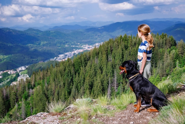 Girl with stands next to her dog of the Rottweiler breed on a peak with vegetation against the cloudy sky