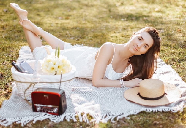 Ragazza con un cappello di paglia in primavera nel parco. bruna con i capelli lunghi si trova su un plaid su uno sfondo di natura estiva. giovinezza e bellezza.