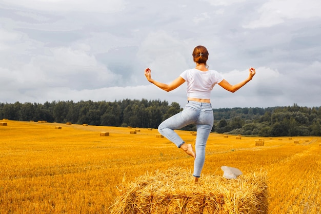 Girl with straw hat stands on a haystack on a bale in the agricultural field after harvesting