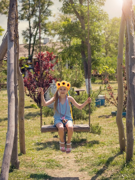 The girl with the sunflowers on her head is swinging on a swing in the village in summer day.