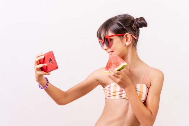 Girl with sunglasses enjoying the summer eating a watermelon making a selfie with the phone White background