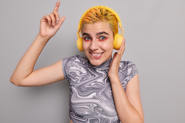 girl with trendy yellow hairstyle bright makeup bites lips catches every rhythm of music wears headphones dressed in casual t shirt on grey