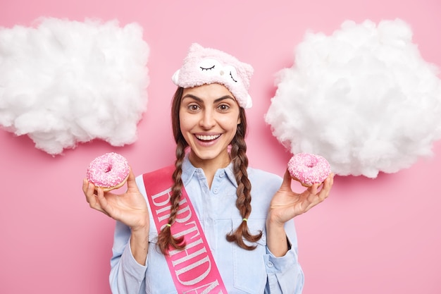 girl with two pigtails smiles positively celebrates friends birthday holds two delicious glazed donuts dressed in casual domestic clothes isolated on pink