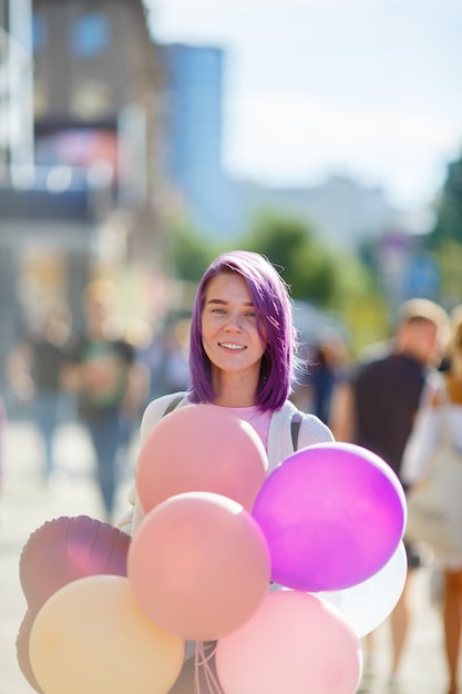 Девушка с фиолетовыми волосами в белом свитере стоит на городской улице с воздушными шариками