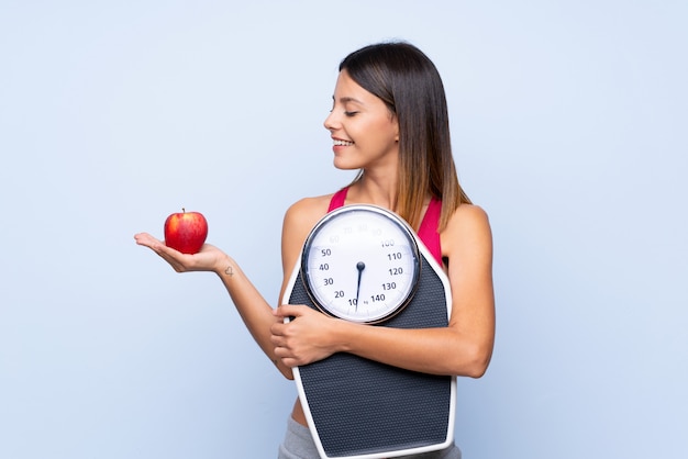 Girl with weighing machine over isolated blue holding a weighing machine while looking an apple