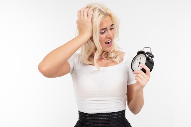 A girl with white hair in a white T-shirt worries that she will be late holding an alarm clock in her hand on an isolated white background