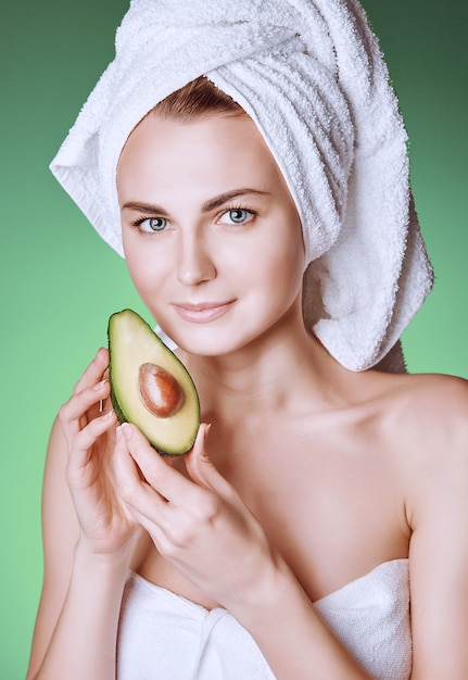 Girl with a white towel on her head with a nutritious green mask on her face and an avocado in her hands on a green background with space for text