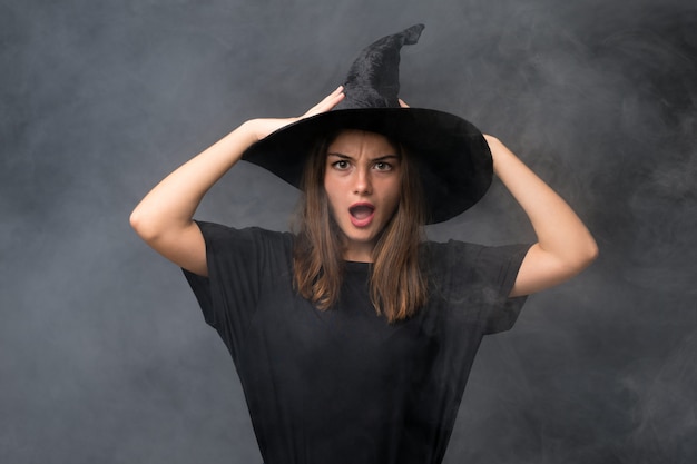 Ragazza con il costume della strega per le feste di halloween sopra la parete scura isolata con espressione facciale di sorpresa