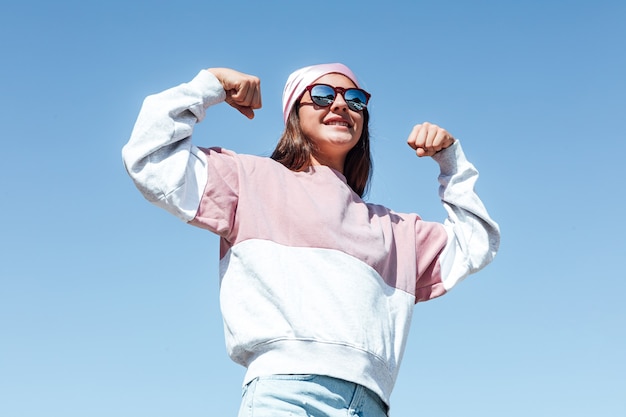 Girl woman with sunglasses and pink head scarf on her head, squeezes her arm as a sign of strength. International Breast Cancer Day, with the sky like background. 