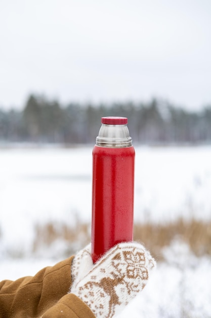 A girl in woolen mittens holds a red thermos with tea in her hands in a winter forest