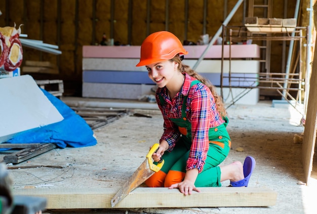 ワークショップで木を扱う女の子木工と木工のコンセプト保護帽子の仕事で木工大工として働くハンドソーで大工を装ったかわいい子供の肖像画