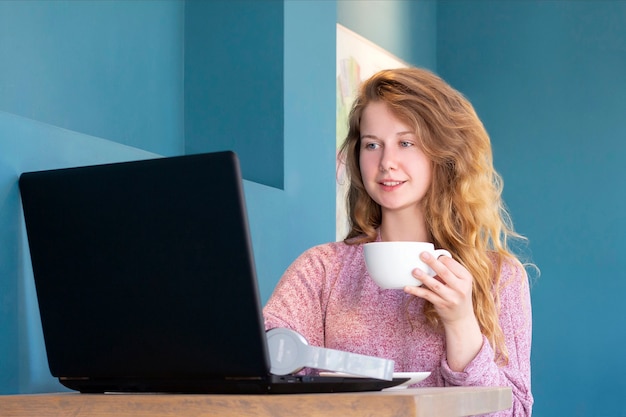 女の子はラップトップで働いています。在宅勤務、オンライン。一杯のコーヒーとカフェの女の子