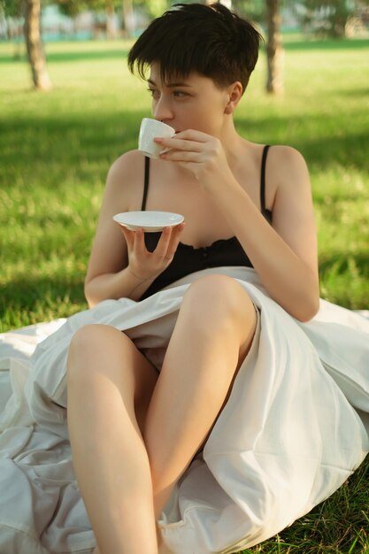 A girl wrapped in a blanket sits on green grass with a cup of coffee enjoying a refreshing morning