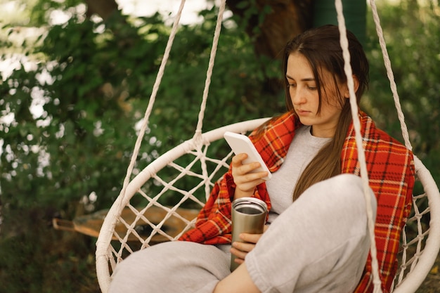 Девушка, завернутая в плед, пьет чай и пользуется телефоном в подвесном кресле на открытом воздухе, люди используют технологии