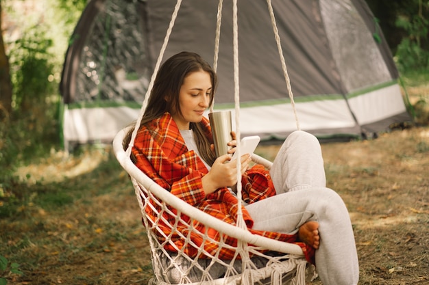 Девушка, завернутая в плед, пьет чай и пользуется телефоном в подвесном кресле на открытом воздухе, люди используют технологии