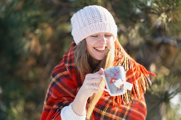 Girl wrapped in a warm plaid drinks winter coffee with marshmallows on the street