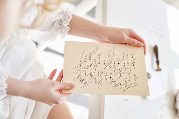 Girl writes a letter to her beloved man, sitting at home at table in a white light dress, purity and innocence. Curly blonde romantic look, beautiful eyes. White wildflowers on the table. Perfect body