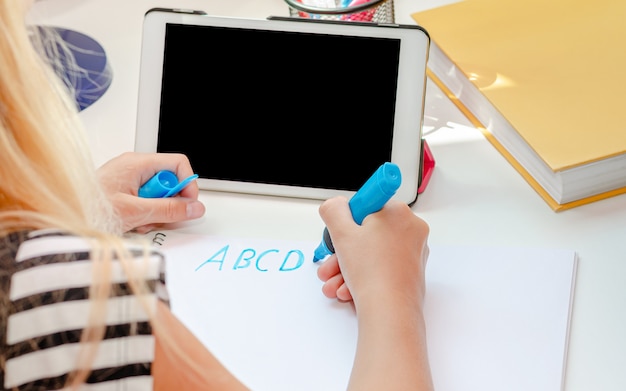 Girl writing letters in the front of tablet with empty black screen. Online English learning concept