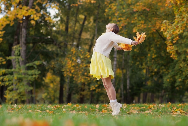 秋の公園で黄色い服を着た女の子は、黄色の葉を両手に持って秋に喜びます