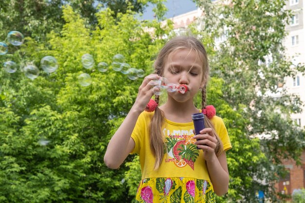 A girl in a yellow dress blows soap bubbles in a park against a background of trees Cute little girl blowing soap bubbles