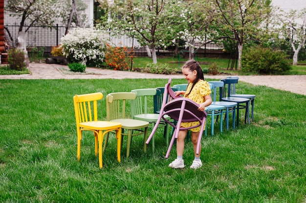 Девушка в желтом платье несет деревянный стул и расставляет стулья в линию, организуя детский ...