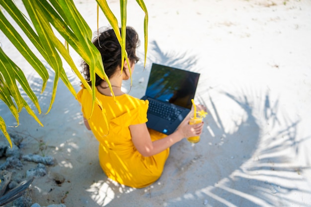 Girl in a yellow dress on a tropical sandy beach works on a laptop and drinks fresh mango. Remote work, successful freelance. Works on vacation.