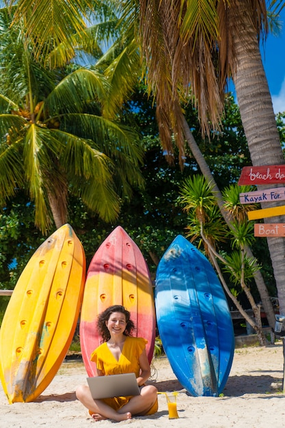 Photo girl in a yellow dress on a tropical sandy beach works on a laptop near kayaks and drinks fresh mango. remote work, successful freelance. works on vacation.