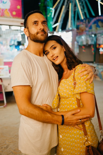 Photo a girl in a yellow dress with a plunging neckline and her boyfriend with a beard are hugging each other between amusement rides. a couple of lovers on a date at the fair in valencia.