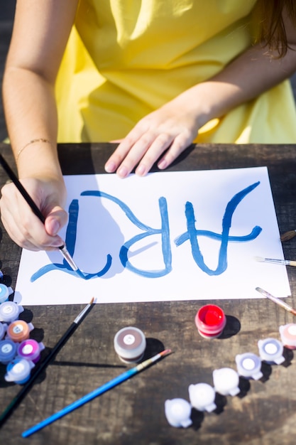 A girl in a yellow dress writes paints on a sheet of paper the word art. 