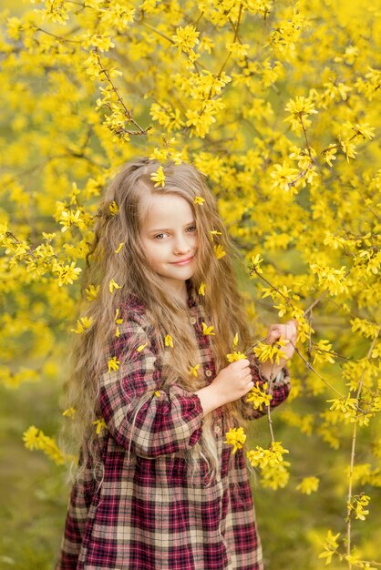 黄色い花の少女。レンギョウを背景にした子供。彼女の髪に花を持つ子供の春の肖像画