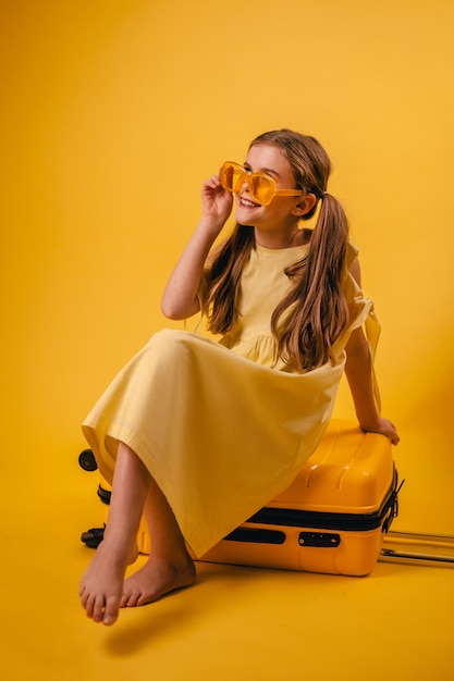 A girl in yellow glasses sits on a suitcase and waits for transport