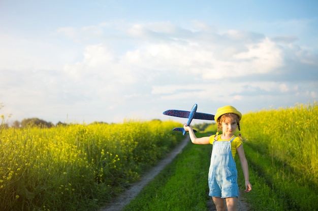 黄色いパナマ帽子をかぶった女の子がおもちゃの飛行機をフィールドに発射します。夏の時間、子供時代、夢と不注意。旅行、冒険、休暇での旅行代理店からの空の旅。村、コテージコア