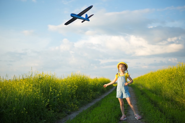 黄色いパナマ帽子をかぶった女の子がおもちゃの飛行機をフィールドに発射します。夏の時間、子供時代、夢と不注意。旅行、冒険、休暇での旅行代理店からの空の旅。村、コテージコア