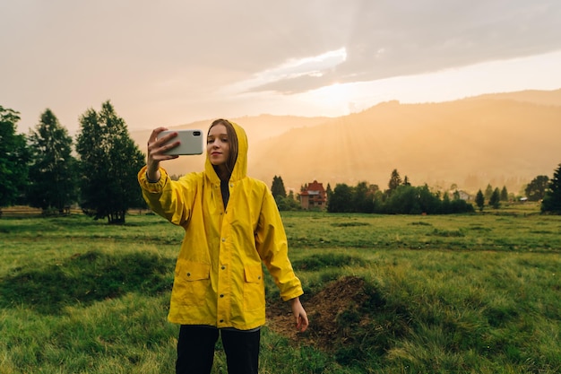 girl in a yellow raincoat takes a selfie on a background of beautiful sunset in the mountains