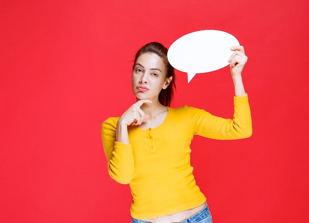 Girl in yellow shirt holding an ovale info board and looks confused and thoughtful