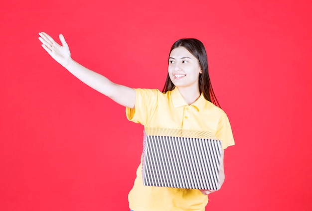 Girl in yellow shirt holding silver gift box and inviting someone to come across.