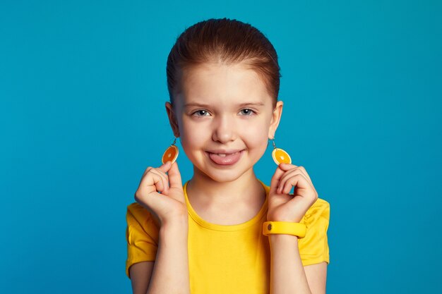 Photo girl in yellow shirt showing tongue and orange earrings against blue wall