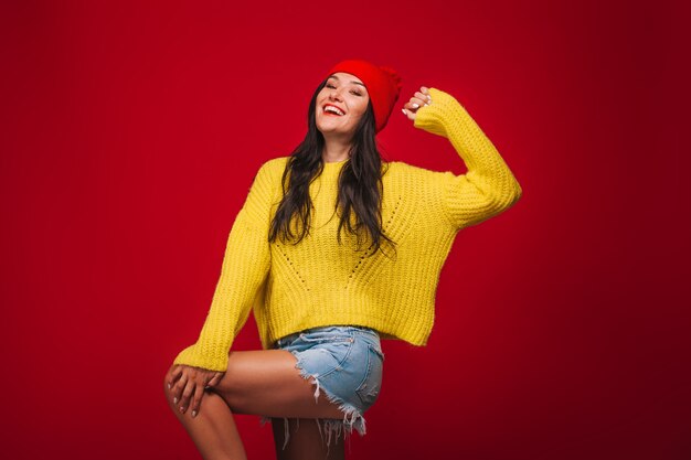 A girl in a yellow sweater and a hat on a red background grimaces