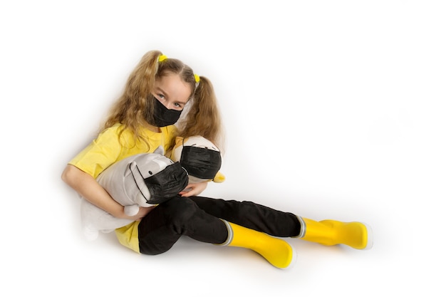 Girl in a yellow t-shirt and black mask covid19 on a white background with toys in a mask. High quality photo