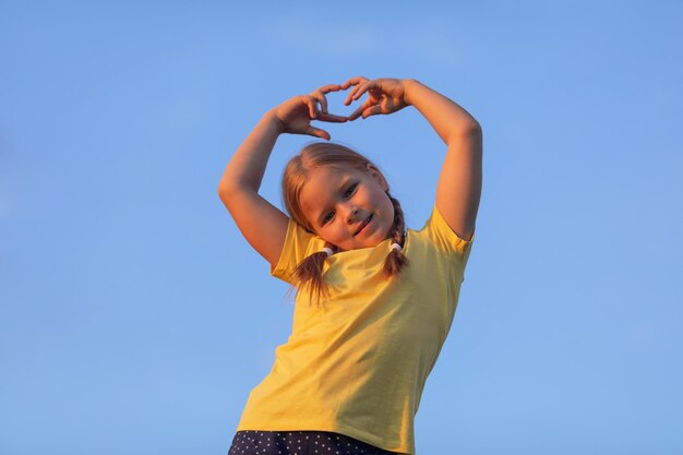 A girl in a yellow T-shirt holds her palms in the shape of a heart against a blue sky at sunset