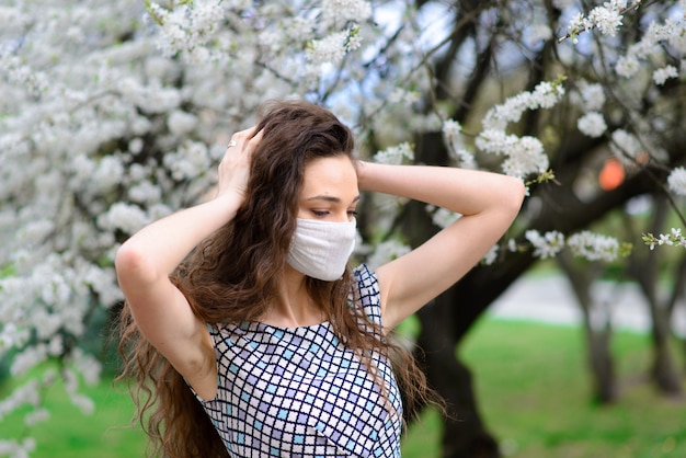 Девушка, молодая женщина в защитной стерильной медицинской маске на лице в саду весной. Загрязнение воздуха, вирус, концепция пандемического коронавируса.