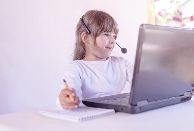 A girlchild in headphones with a microphone and a laptop communicates online at home